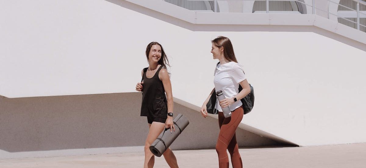 Two women in active wear walking outdoors while smiling and carrying yoga mats.
